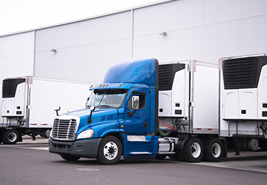 A semi-truck with a reefer trailer attached, exiting a warehouse parking lot.