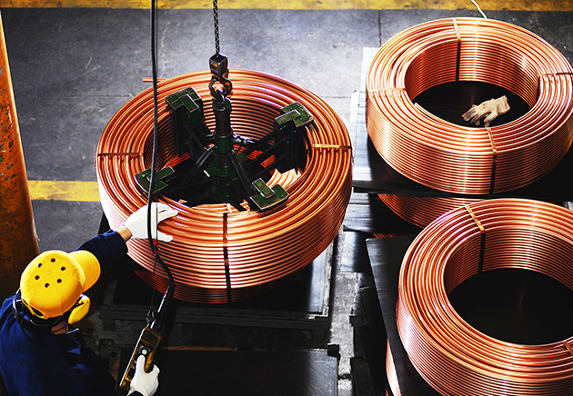 Worker using a mechanical lift on large spool of copper coil with two more copper coils on the side.