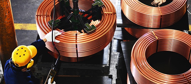 Worker using a mechanical lift on large spool of copper coil with two more copper coils on the side.