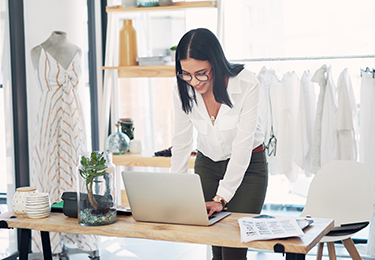 A female business owner standing over a desk, using her laptop at her clothing store.