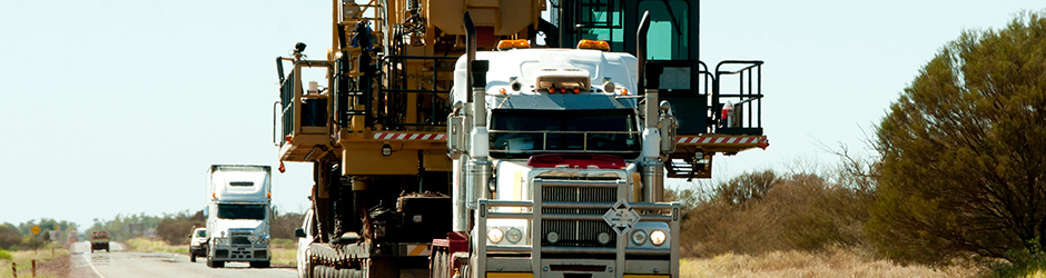 A white truck carrying a wide oversized cargo of heavy machinery.