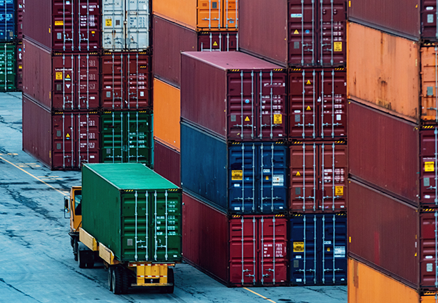 A truck transporting a shipping container with stacks of shipping containers nearby.