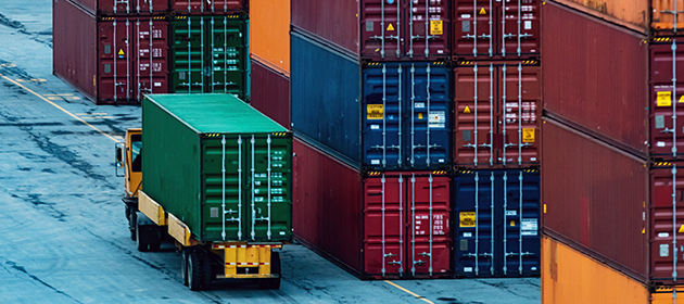 A truck transporting a shipping container with stacks of shipping containers nearby.