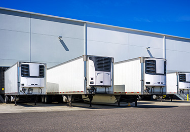 A row of four white refrigerated trailers parked outside a warehouse.